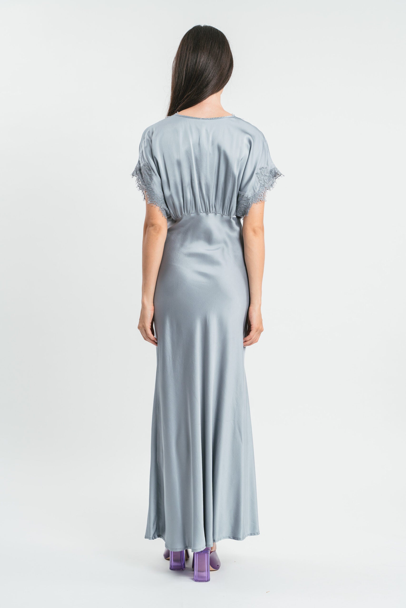 Modella indossa abito argento lungo in viscosa elasticizzata con rifiniture in pizzo francese. Retro